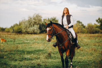 Young beautiful blonde woman jockey rides a brown horse in a meadow at sunset in summer. Preparing for an equestrian competition.
