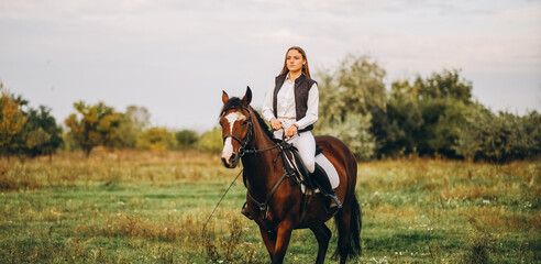 Young beautiful blonde woman jockey rides a brown horse in a meadow at sunset in summer. Preparing...