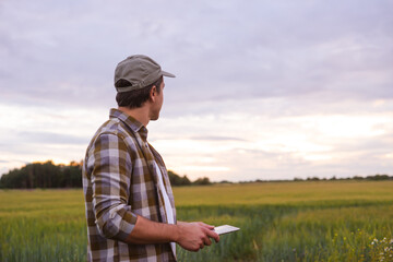 Farmer with a tablet computer in front of a sunset agricultural landscape. Countryside field. The concept of country life, food production, farming and technology concept.