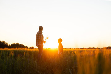Fototapeta na wymiar Farmer and his son in front of a sunset agricultural landscape. Man and a boy in a countryside field. Fatherhood, country life, farming and country lifestyle concept.