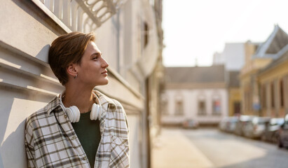 Short red hair woman standing on street with headphones on her neck leaning on wall building.