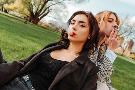 Two pretty women friends sitting on grass smoking cigarette. Couple of gay lesbian girls hugging embracing together girlfriends, have a date. LGBT concept. Addiction, cancer, tobacco. Bad habit
