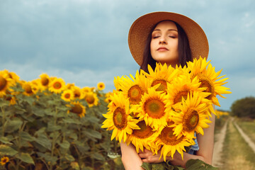 Lovely woman hold yellow bouquet bunch blooming sunflower field outdoors sunrise warm nature background. Lady dressed blue dress wear hat posing standing outside, agriculture concept
