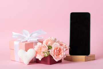 Smartphone on the background of a pink gift box with roses and a knitted white heart on a light pink background with a place for text.