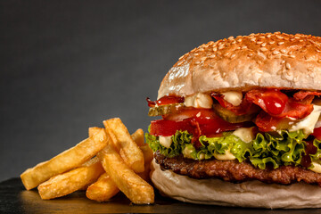 Bacon hamburger with onions tomatoes cucumber lettuce and french fries against dark gray background.