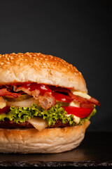 Bacon hamburger with onions tomatoes cucumber and lettuce against dark background. Abstract fast food photo.