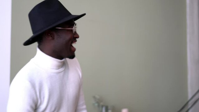 Cheerful hipster model in black hat enjoying time during leisure pastime indoors, handsome African American male in classic eyeglasses talking and laughing in stylish fashion apparel