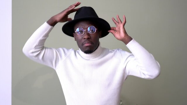 Emotional dark skinned male model dressed in trendy white wear dancing and having fun during positive daytime indoors, happy African American hipster guy in optical spectacles and black hat