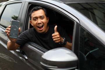 Happy asian driver pointing his cell phone screen and showing thumbs up in his car