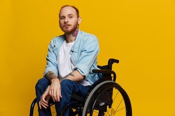 A man in a wheelchair looks at the camera, with tattoos on his arms sits on a yellow studio background, the concept of health is a person with disabilities