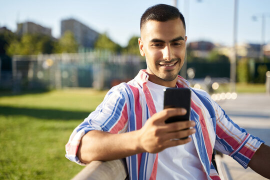 Young smiling Iranian man smiling happy using smartphone at the city. Handsome young guy dressed in casual clothes relaxing outdoors at the city street