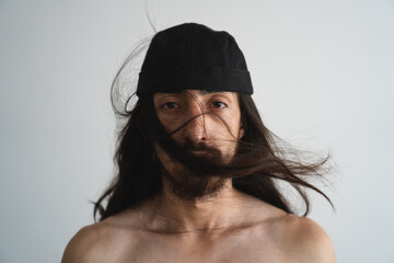 Young Latin man with long hair and no shirt wearing a beanie looking inexpressive at camera with a...