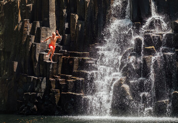 Middle-aged man jumping in waterfall lake. Falling water streams flow on black volcanic stone...