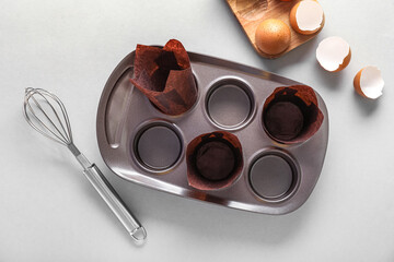 Muffin baking tray with paper forms, whisk and eggs on light background