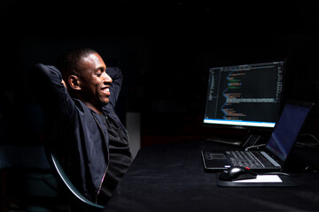Black programmer smiling while sitting back looking at code in a dark room