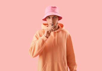 Young man in bucket hat smoking disposable electronic cigarette on pink background