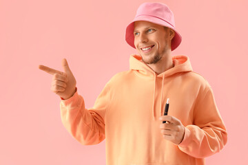 Young man in bucket hat with disposable electronic cigarette pointing at something on pink...