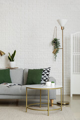 Interior of living room with grey sofa, lamps, houseplants and coffee table near white brick wall