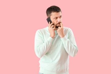 Handsome man biting nails and talking by mobile phone on pink background