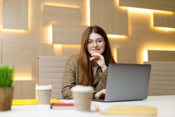 Positive woman in the workplace, a young employee sits at a table with a laptop.