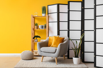 Interior of living room with folding screen, armchair and houseplant near yellow wall