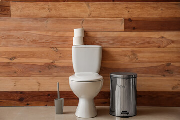 Toilet bowl with paper rolls, brush and bin near wooden wall