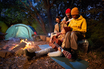 Multiracial colleagues sitting together beside fire stake playing on the guitar discussing and singing. Group of friends with guitar near bonfire and camping tent outdoors overnight. Copy space.