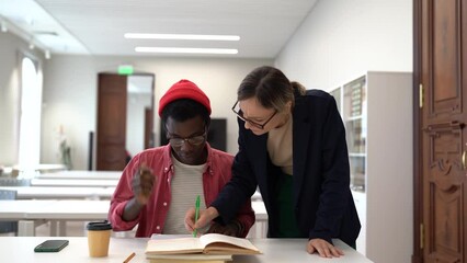 Female teacher helping University student to improve grades and academic performance, young African guy preparing for exam with private tutor in library. Tutoring services and education concept