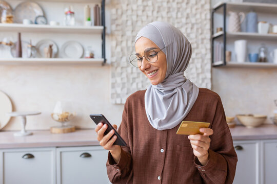 Beautiful muslim girl close up in kitchen, woman in hijab smiling and happy holding smartphone and gold bank credit card, shopping online in online store at home.