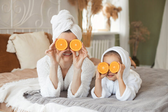 Funny beautiful mother and cute little daughter lying on bed and playing with fresh fruit orange for face mask having spa day toghether at home