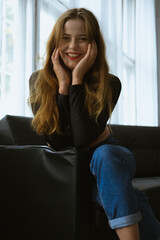 Brunette girl in the blue jeans is sittieng  on a black sofa and smileing 