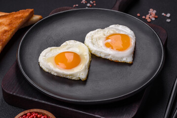 Two heart-shaped fried eggs on a black ceramic plate on a dark concrete background