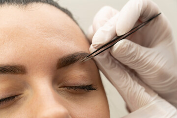 close-up of a brunette woman makes shape and paint eyebrows with tweezers in a beauty salon.