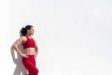 Sporty woman with hands on her hips leaning against a white wall in the sun.