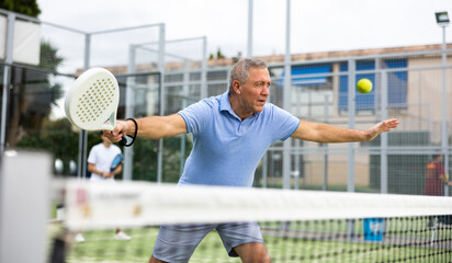 Emotional mature male playing padel behind the net during match on tennis court in autumn