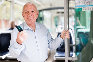 Senior European man with smartphone in hand standing inside tram and waiting for his stop.