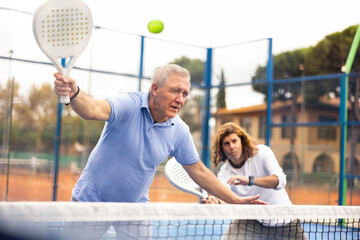 Sporty mature man padel player hitting ball with a racket on a hard court - obrazy, fototapety, plakaty