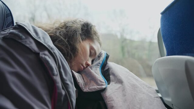 Woman sleeping on a moving bus wearing warm a coat as it drives on a highway. Tired and exhausted female traveler falls asleep on a public bus ride. Her head is resting on a coat