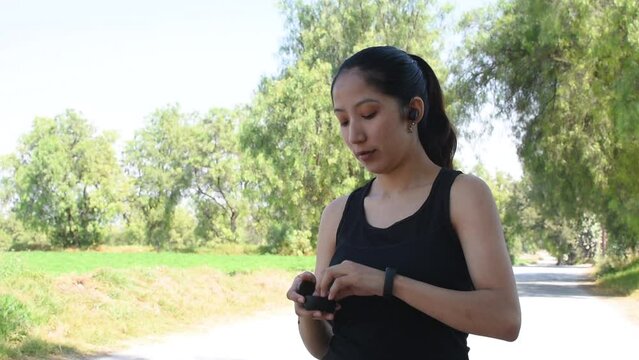 Joven mujer caucasica haciendo ejercicio y colocandose sus audifonos inalambricos antes de salir a correr en la naturaleza video en HD 