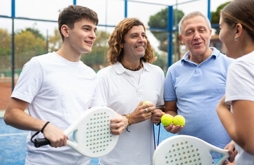 Young woman, guy and two mature men holding rackets and balls chatting friendly after paddle tennis...