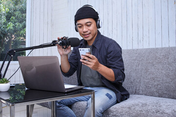 Asian man making a podcast from home studio while holding mobile phone. Recording a podcast for...
