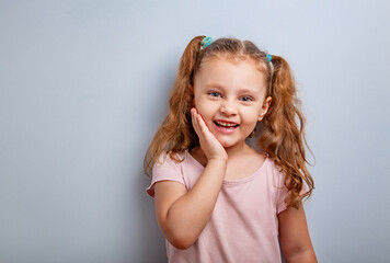 Emotional fun kid girl smiling with hand at face and looking happy with ponytail hairstyle on blue...