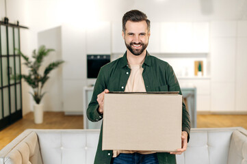 Positive handsome stylish caucasian man, stand at home in living room, holding a large cardboard box, received parcel from the online store, preparing to unpack, looks at camera, smile, happy emotions