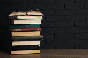 Stack of old hardcover books on wooden table near black brick wall, space for text