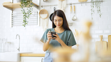 Happy young asian woman relaxing at home. Asia female standing at counter kitchen and using mobile smartphone