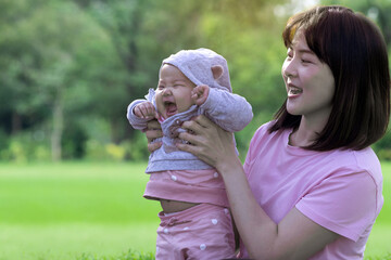 Obraz premium Asian mother carry funny baby girl, relax time on green lawn, cute baby laughing happily, happy family Asian mom and her daughter playing in the outdoor park