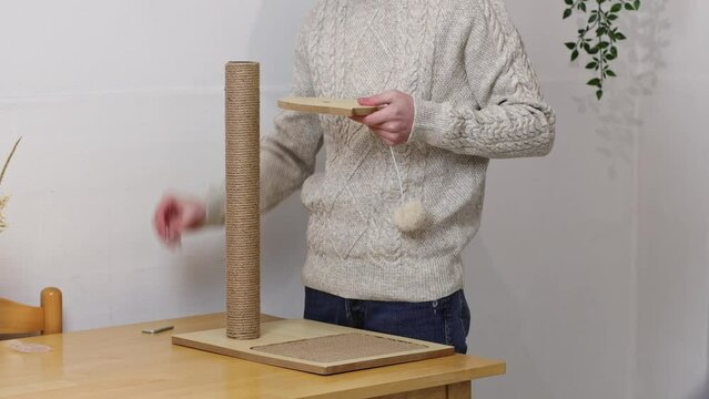 Young caucasian man in a knitted sweater assembles a creative wooden scratching post for a cat by manually twisting a screw while standing at a table in the room, close-up side view.Concept of assembl