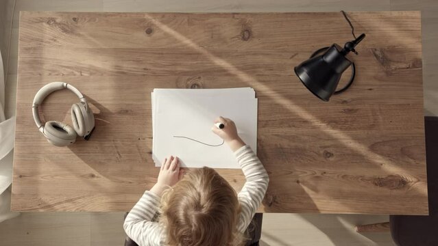 little blonde girl starts drawing with black marker, top down view of child at work desk at home or in kindergarten. Kid draws doodles on white blank sheet.