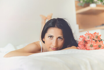 Obraz na płótnie Canvas a beautiful young woman is lying in bed, looking at the camera and smiling, pink flowers in the foreground, selective focus