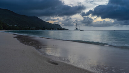 A serene evening on a tropical island. Ocean waves roll onto the beach. Wet sand glistens. Yachts...